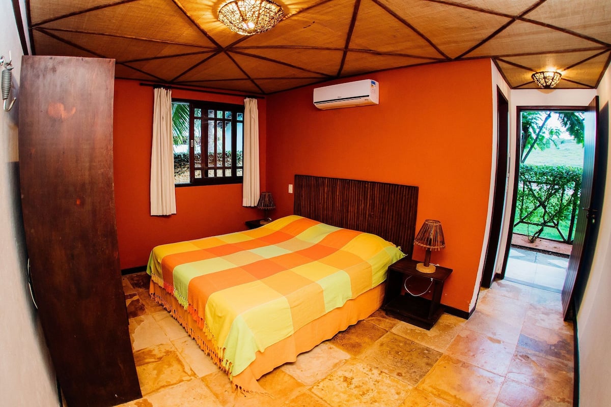 A bedroom with a bed, an air conditioner, a nightstand, a lamp, and a view of greenery outside.