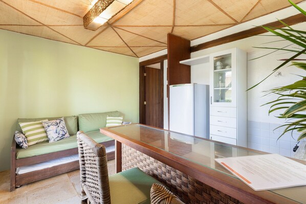 Una habitación con una mesa de vidrio, un sofá con cojines, una silla de mimbre y un refrigerador.