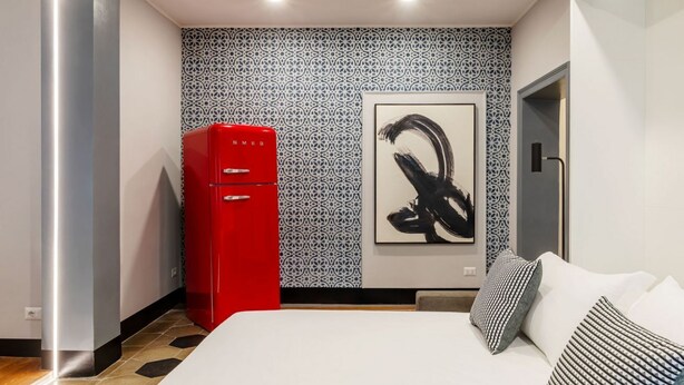 Ein modernes Hotelzimmer mit einem Bett, einer roten Minibar, strukturiertem Wandpapier und einem schwarzen abstrakten Gemälde.