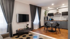Ein modernes Wohnzimmer mit Flachbildfernseher, einem Essbereich mit rundem Tisch und Stühlen sowie einer Küche mit Mikrowelle und Pflanze.