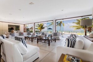 A spacious living room with white sofas, striped pillows, and a large window offering a view of the ocean and palm trees. - Casa dos Corais - Foot in the Sand by Carpediem (Ceara-Mirim)