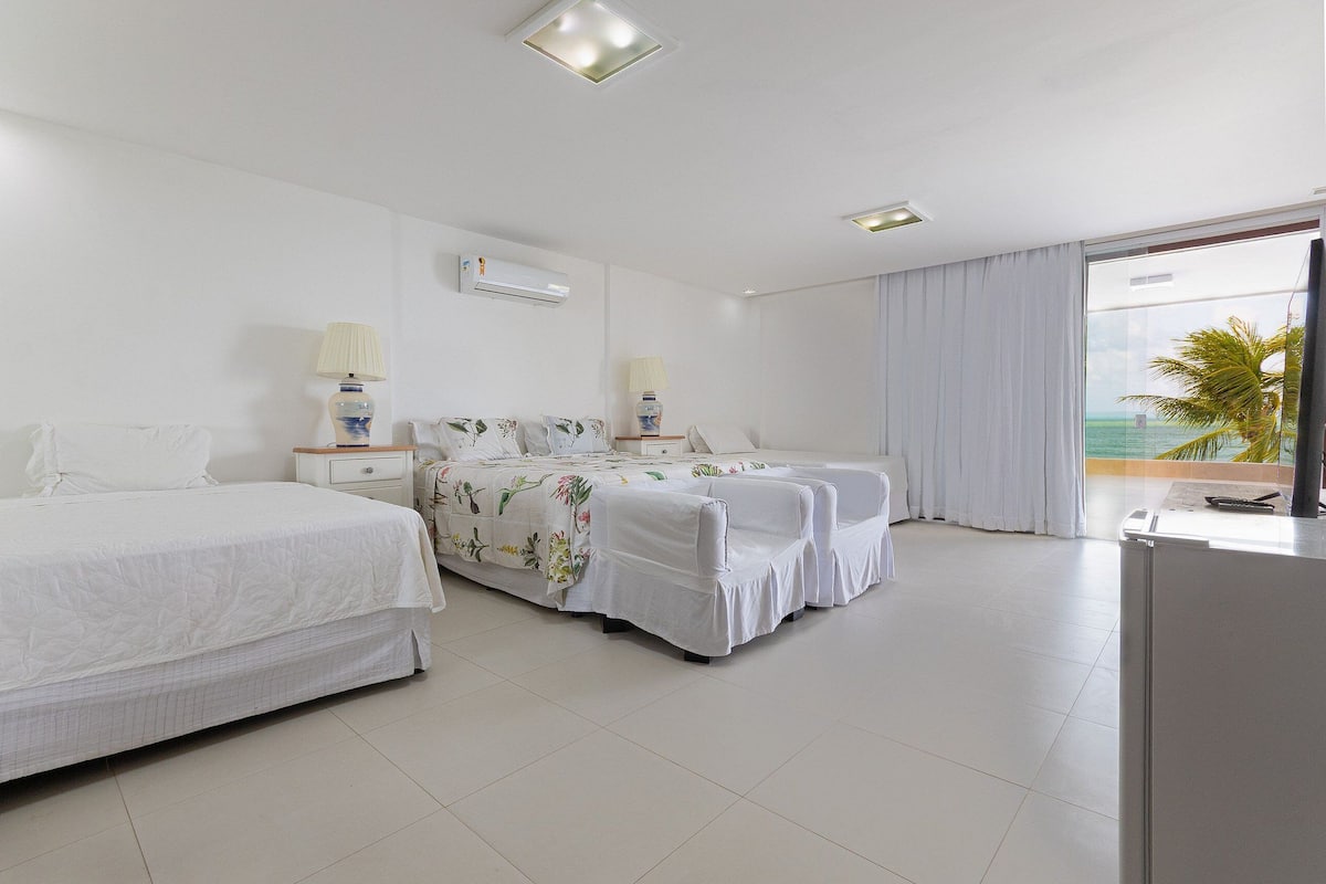 A hotel room with two beds, a large window with a view of the ocean, and a balcony.