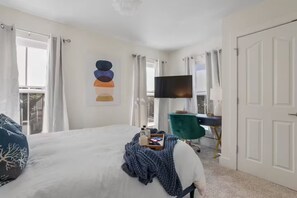 A bedroom with a large bed, a desk with a TV, and a window with curtains. - Boutique Downtown Cottage with Golf Cart (Ocean Springs)