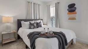 Een keurig opgeruimde slaapkamer met een bed, nachtkastje, lamp en een lijst met een kunstwerk aan de muur.
