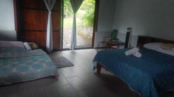 A room with two beds, a small table, and a view of greenery outside.