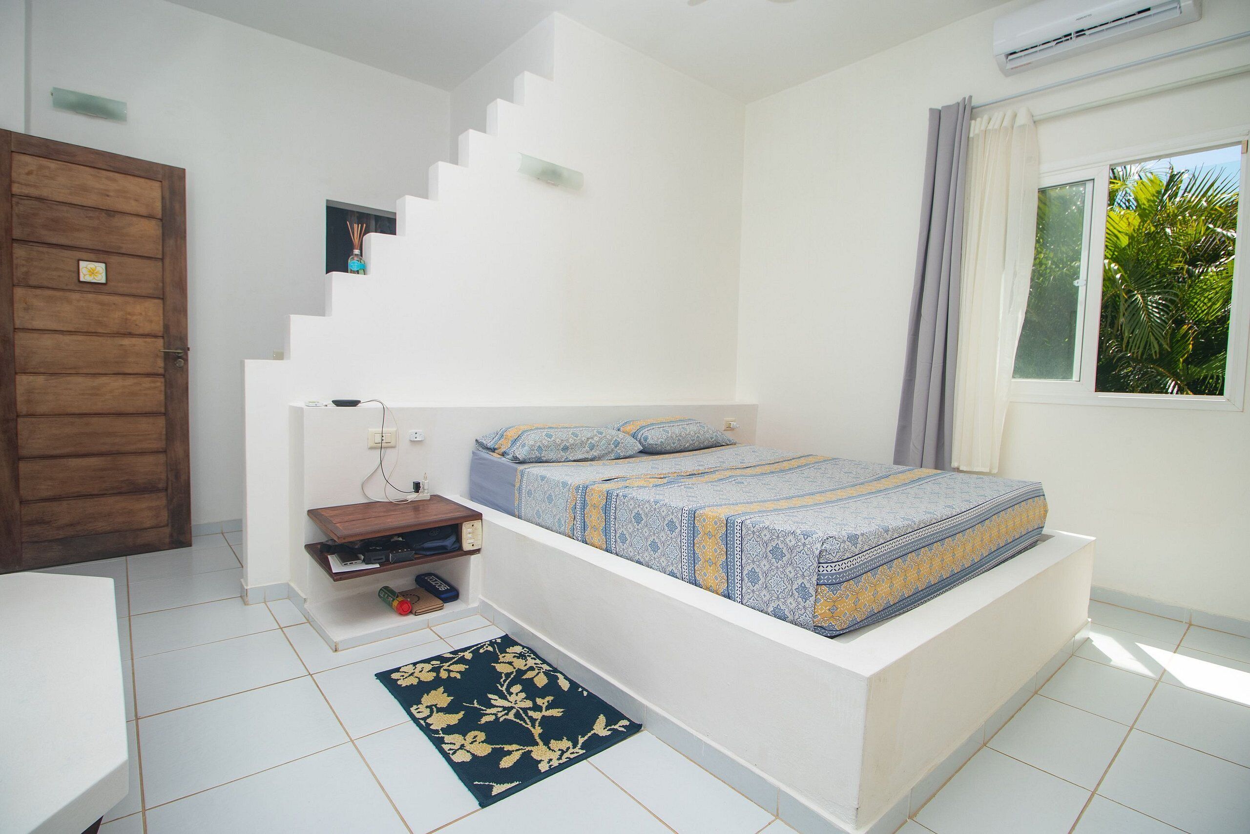 Un dormitorio con cama, una puerta de madera, un estante pequeño, una ventana con cortinas y una escalera.