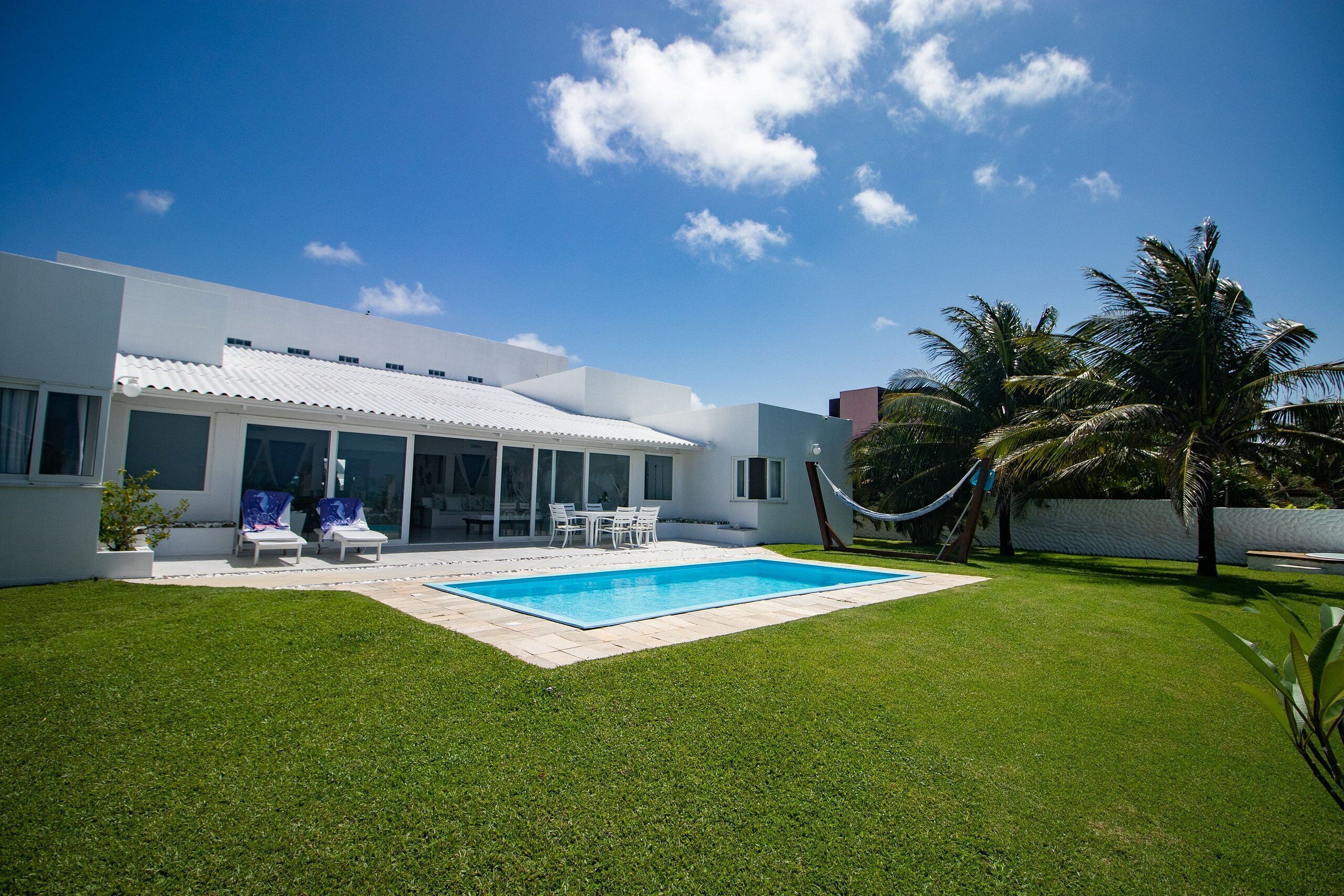 A modern house with a swimming pool, a patio with chairs, and palm trees.