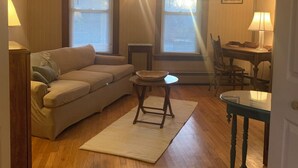 A living room with a sofa, a small table, a lamp, and a window with blinds. - Cozy 1-bedroom apartment in old farm house in historic South Lee (Lee)