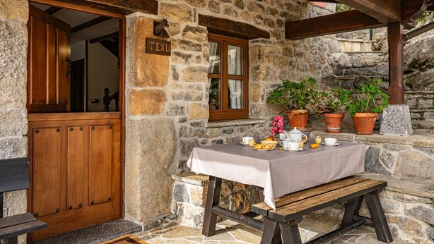 Ein Steinhaus mit einer Holztür, auf der 'TEXU' steht, einem Tisch mit Tischdecke, Topfpflanzen und einer Bank davor.