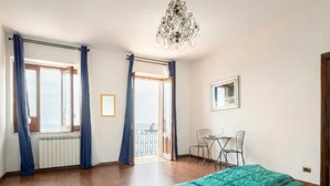 A bedroom with a bed, a chandelier, a window with curtains, a small table with chairs, and a view of the outdoors. - Il Pescatore Elegant Apartment with Stunning Balcony on Isola Bella, Taormina (Mazzarò)