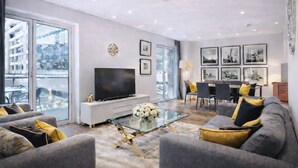 Een moderne woonkamer met een flatscreen televisie, grijze banken met gele kussens, een glazen salontafel en een eethoek met een tafel en stoelen.