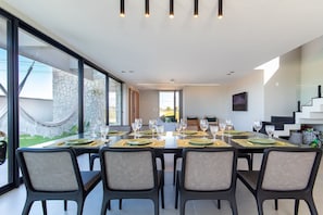 Moderni ruokailutila, jossa on pitkä ruokapöytä kuudelle henkilölle, pöydän ympärillä tuoleja ja näkymä kiviseinälle sekä ulkona oleva riippumatto.