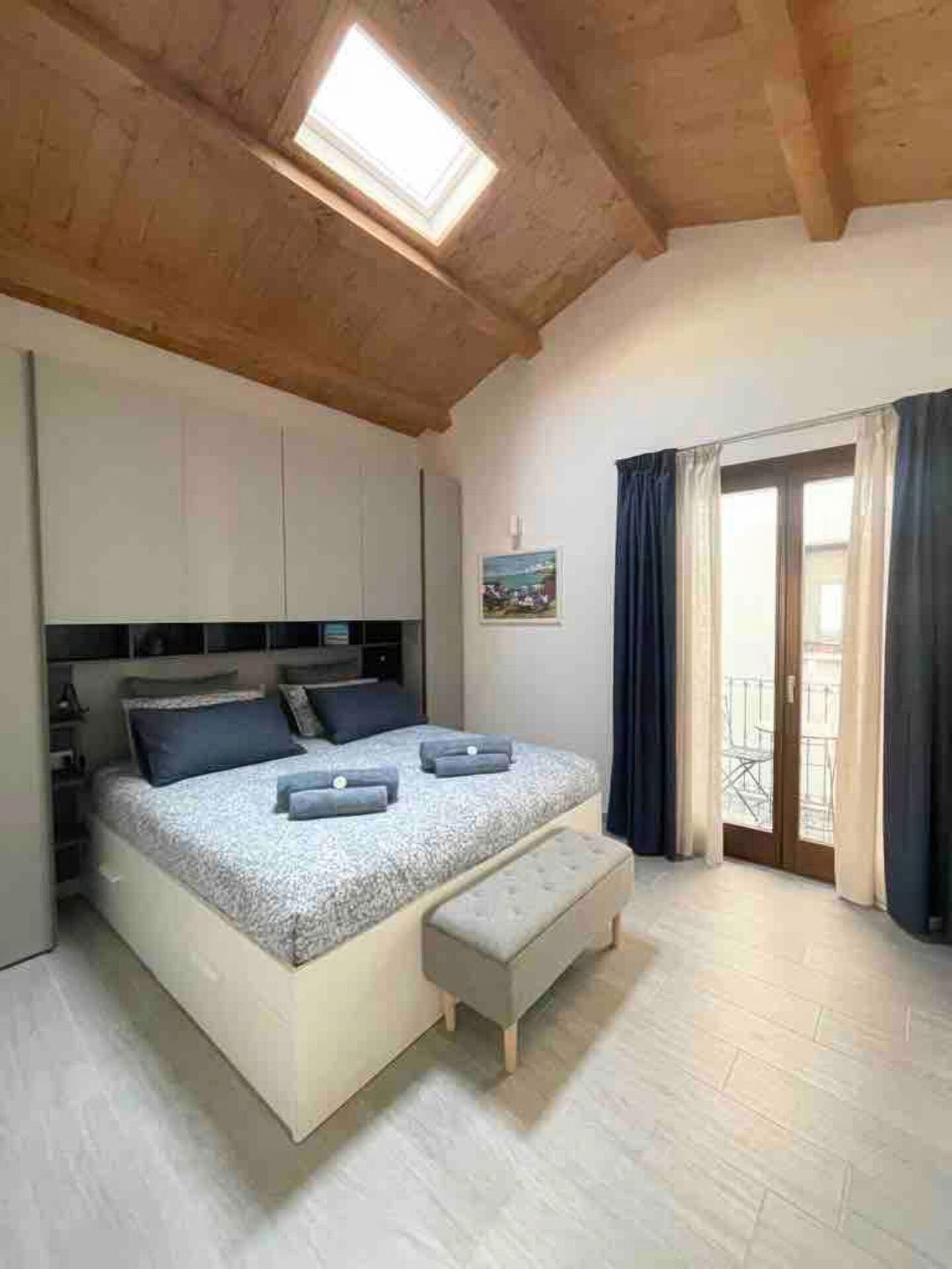 A bedroom with a bed, a bench, a skylight, a door with a view, and a painting on the wall.
