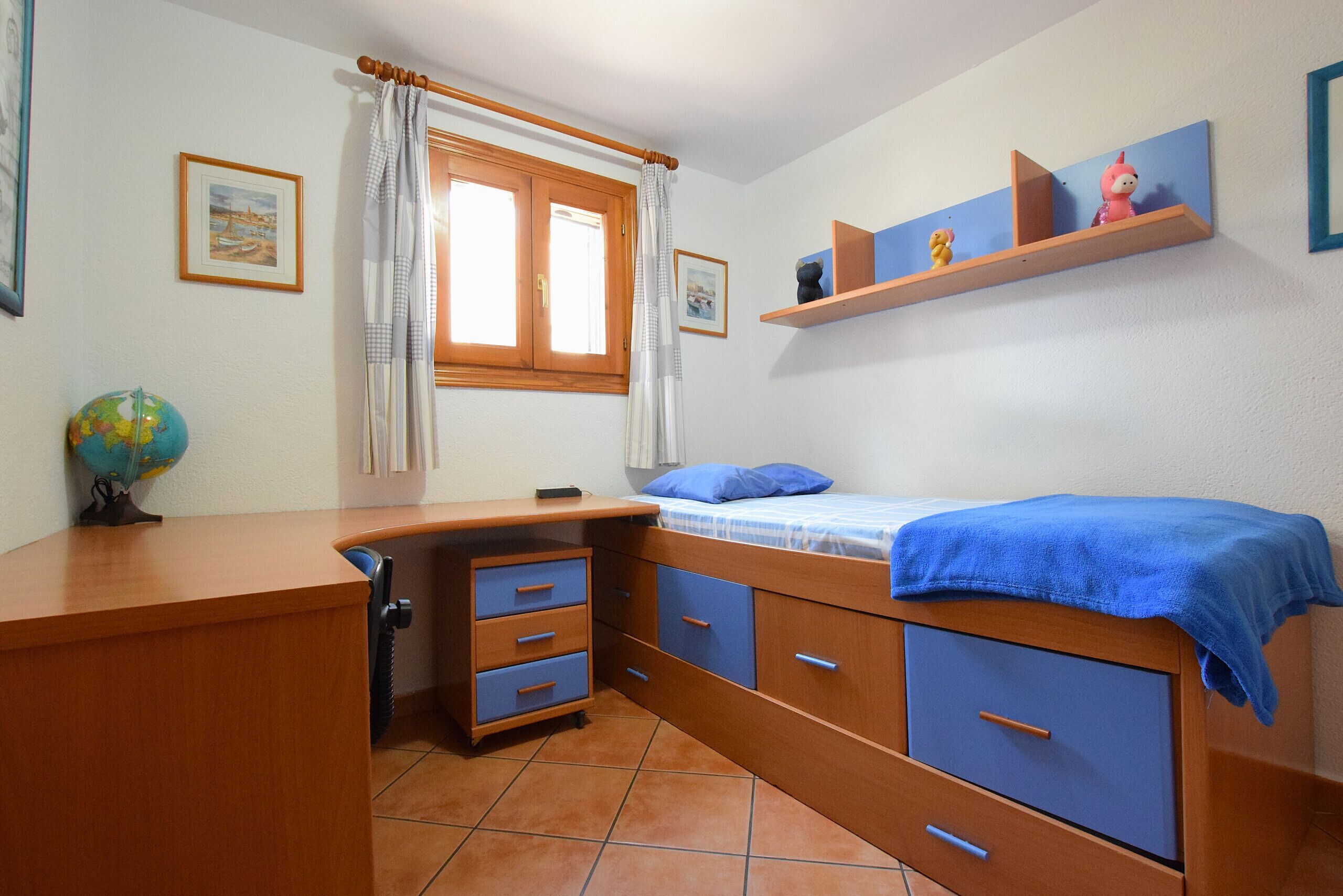 Un dormitorio con un escritorio de madera, una cama con manta azul, un globo terráqueo y una ventana con cortinas.