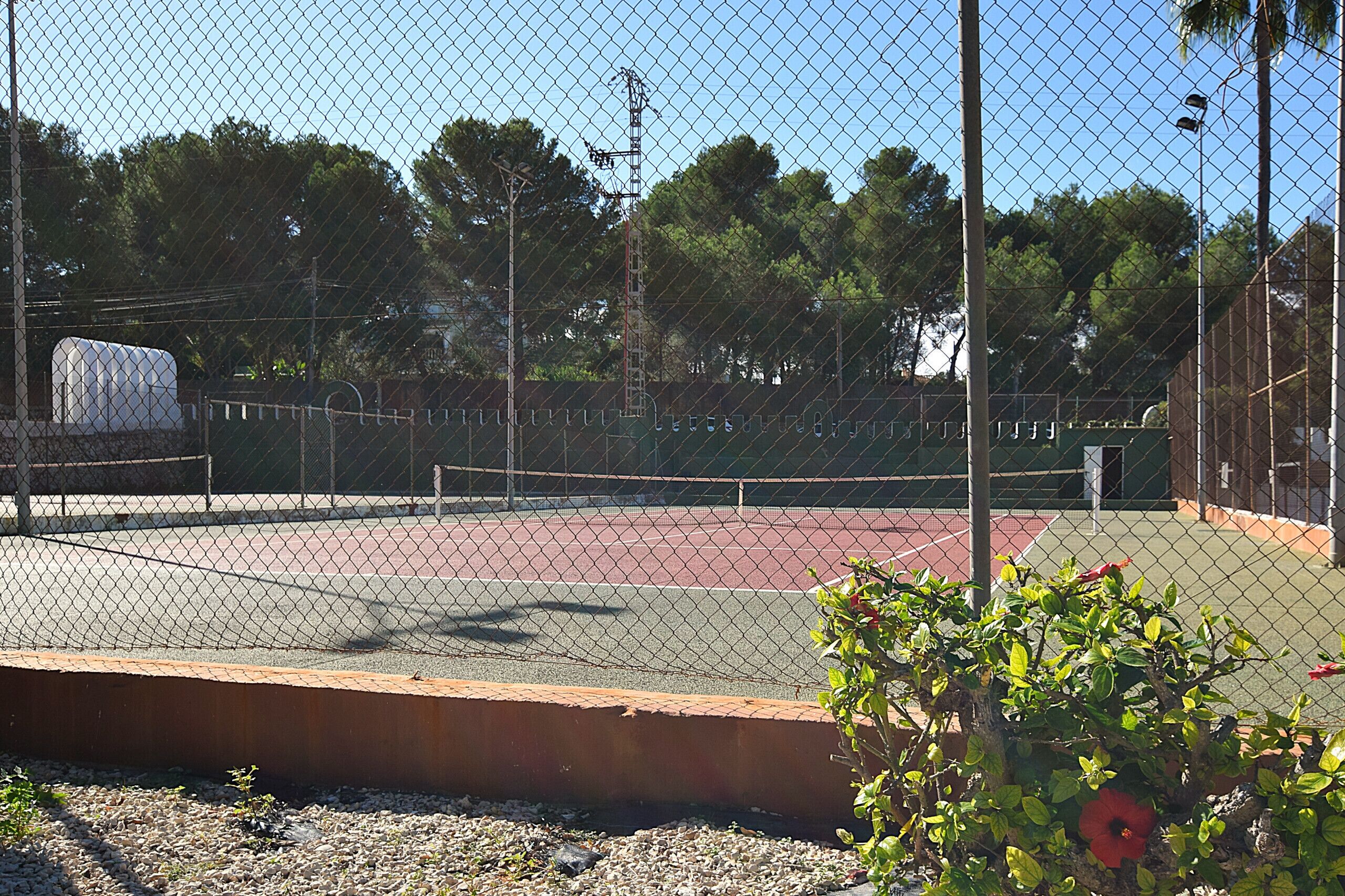 Un campo da tennis recintato con cielo sereno, alberi e un edificio sullo sfondo.