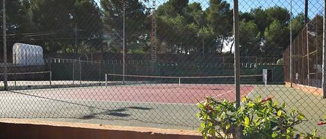 Un campo da tennis recintato con cielo sereno, alberi e un edificio sullo sfondo.