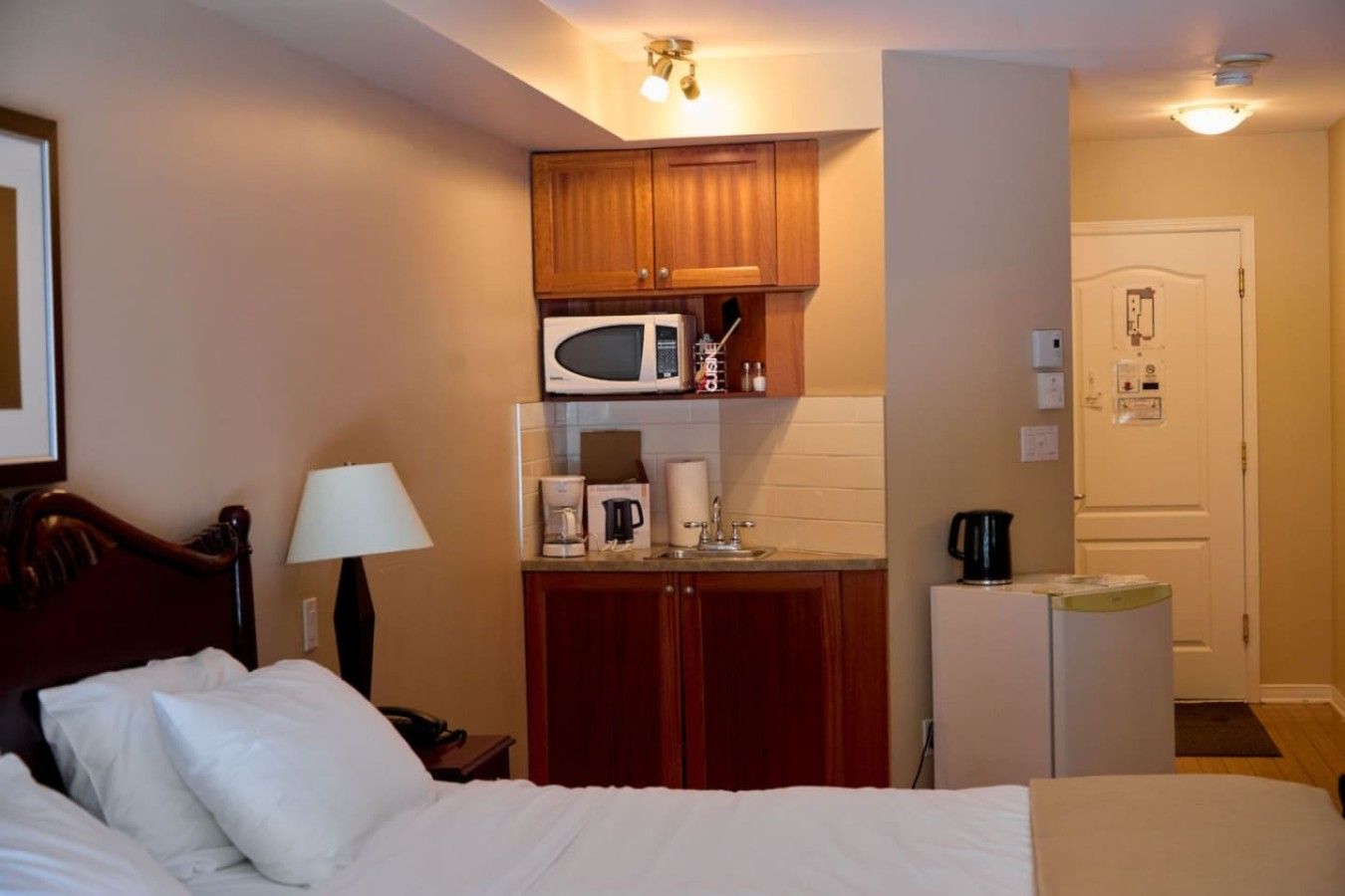 A hotel room with a bed, a bedside lamp, a microwave, a coffee maker, and a refrigerator.
