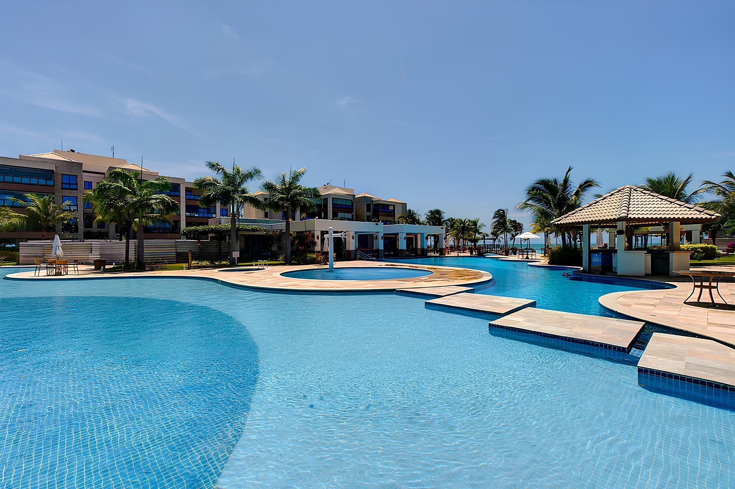A large, multi-level swimming pool with a circular design, surrounded by palm trees and modern buildings.