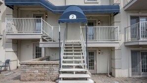 Un immeuble à plusieurs étages, avec un escalier menant à l’entrée, un espace couvert portant le numéro « 2 », et un banc en pierre.