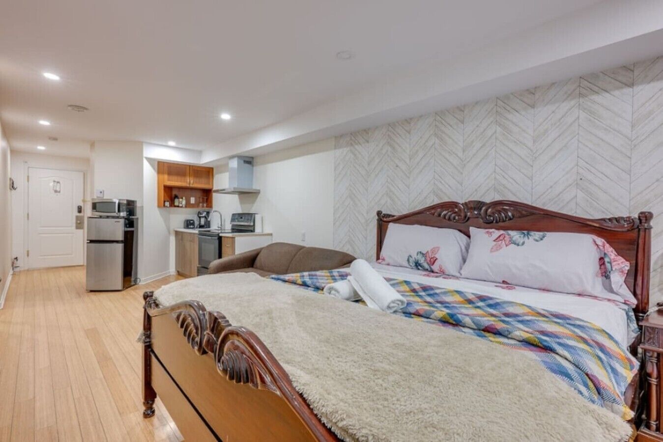 A bedroom with a wooden bed, a small kitchenette, and a herringbone-patterned wall.