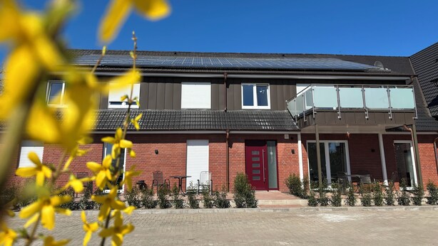 A two-story building with a red door, solar panels on the roof, and a small outdoor seating area with tables and chairs. - Vacation apartment 6 persons, near Cloppenburg, bedroom with TV and single beds (Cappeln (Oldenburg))