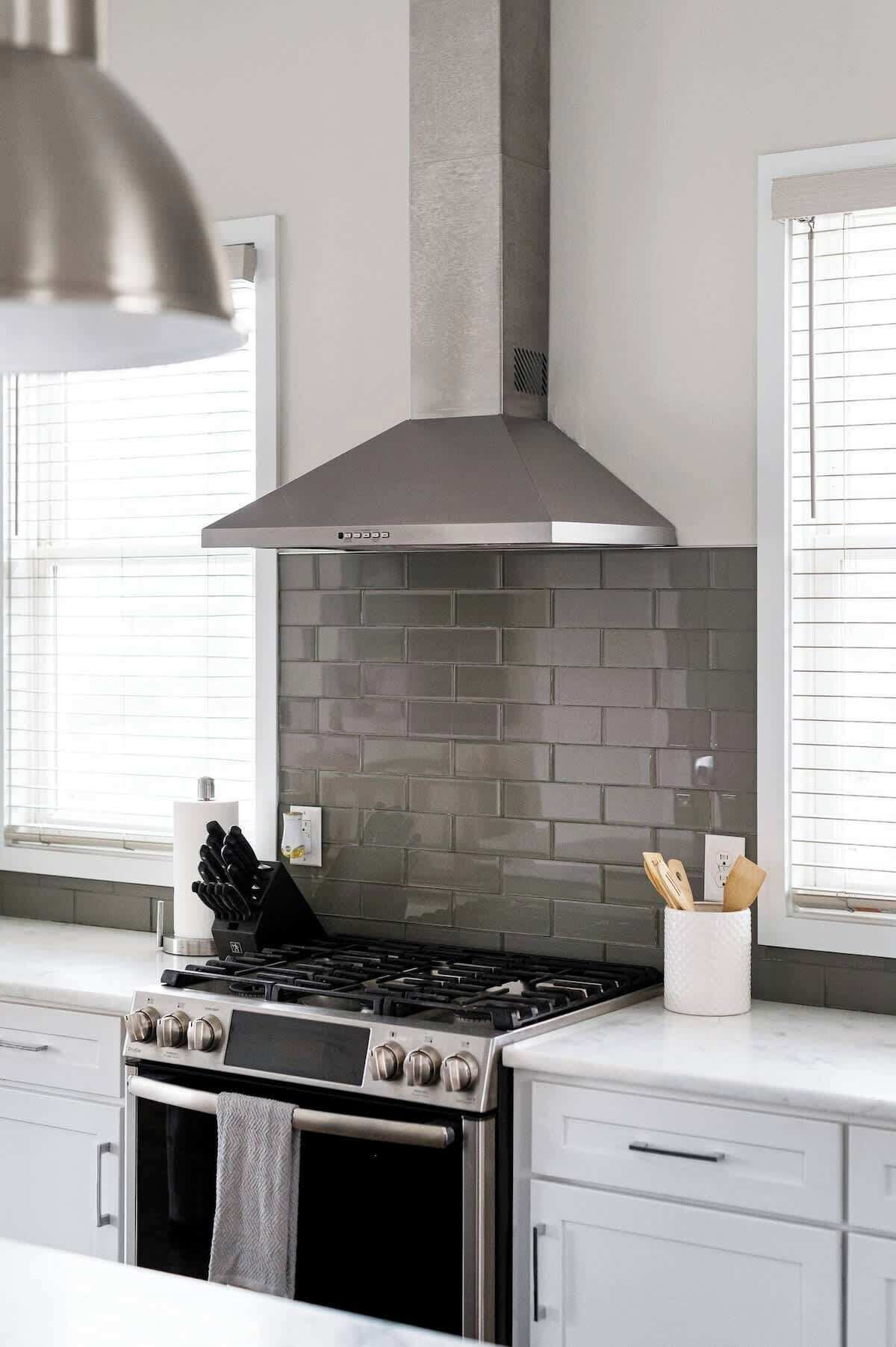 A modern kitchen with a stainless steel stove, a grey brick backsplash, and a tall range hood.