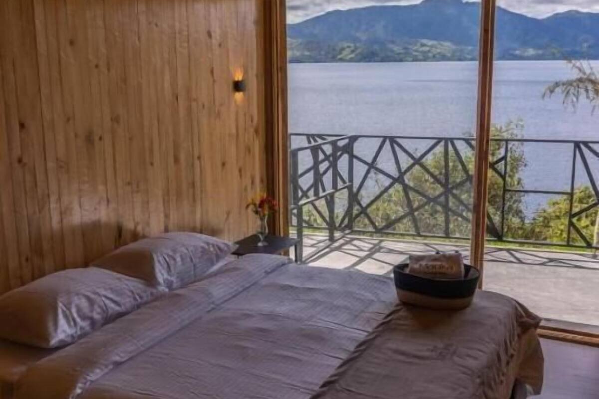 A wooden-paneled room with two beds, a view of a lake and mountains, and a balcony with a railing.