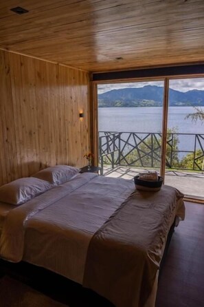 A wooden-paneled room with two beds, a view of a lake and mountains, and a balcony with a railing. - ECOHOTEL MAUKA (San Juan de Pasto)