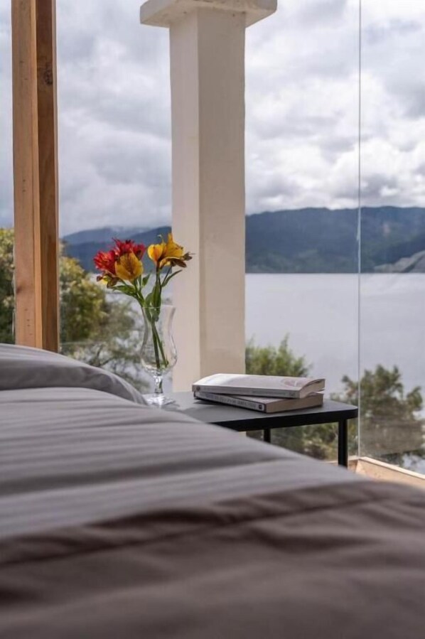 A bed with a view of a lake and mountains, a vase with flowers, and a stack of books on a nightstand.