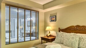 A hotel room with a bed, a bedside table with a lamp, a window with blinds, and a picture on the wall. - Condo Alpina – 3105 | Cozy Retreat in St-Sauveur (Piedmont)