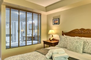 Quarto de hotel com cama, mesa de cabeceira com luminária, janela com persianas e um quadro na parede.