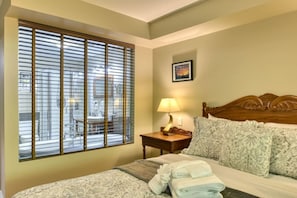 A hotel room with a bed, a nightstand with a lamp, a window with blinds, and a picture on the wall.