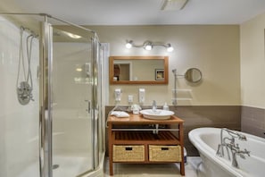 A bathroom with a glass shower enclosure, a wooden vanity with two sinks, a bathtub, and a mirror with lights above.