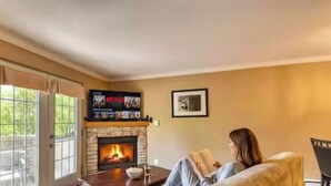 A living room with a fireplace, a television mounted above it, and a person reading a book. - Condo Alpina – 3105 | Cozy Retreat in St-Sauveur (Piedmont)