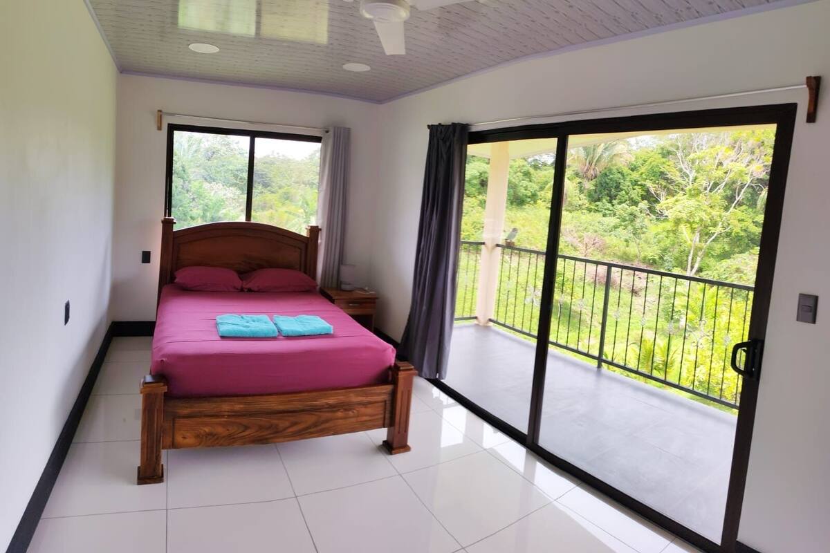 Een slaapkamer met een houten bed, een plafondventilator, een groot raam en een balkon met uitzicht op groen.