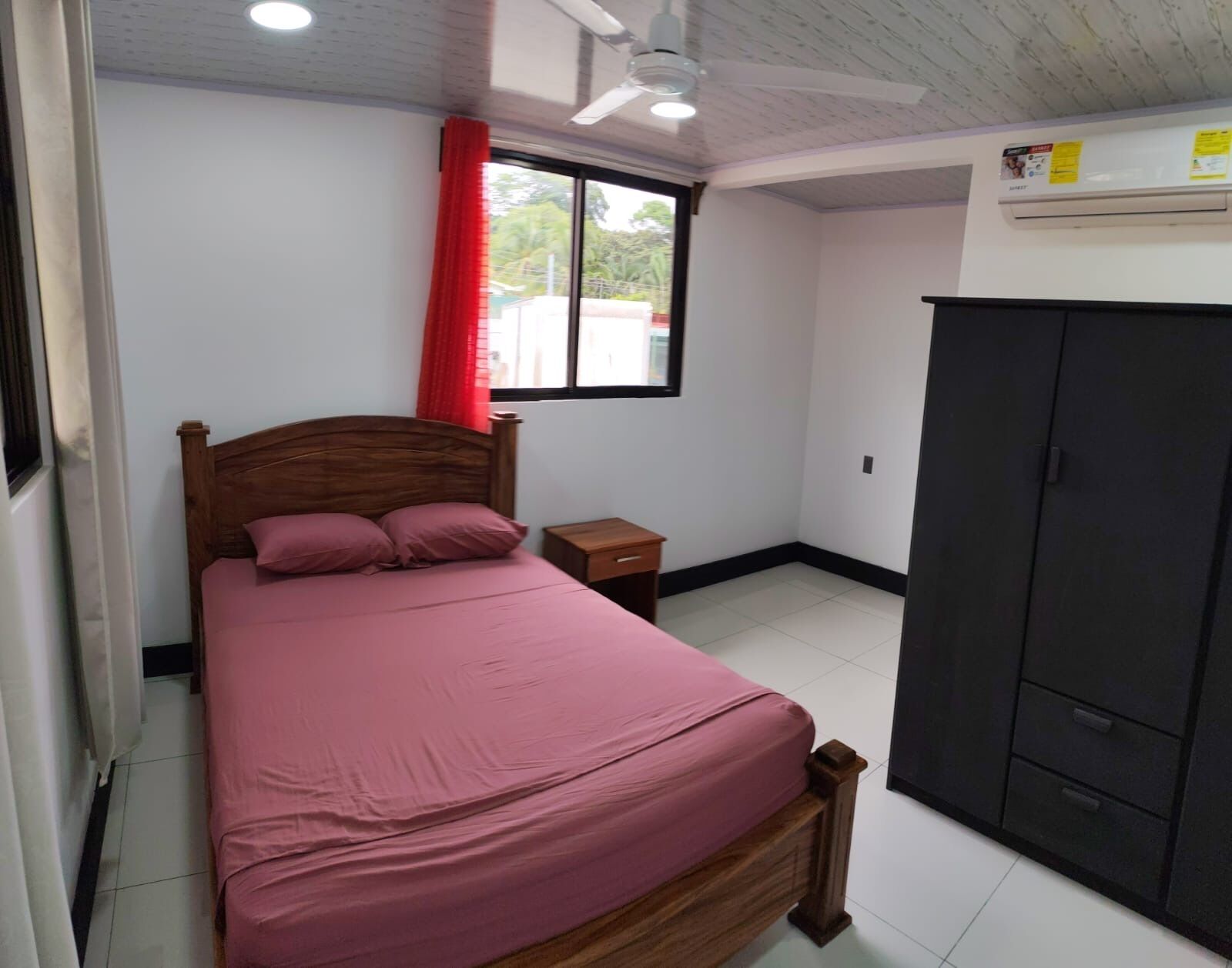 A bedroom with a bed, a nightstand, a wardrobe, a window with red curtains, and a ceiling fan.