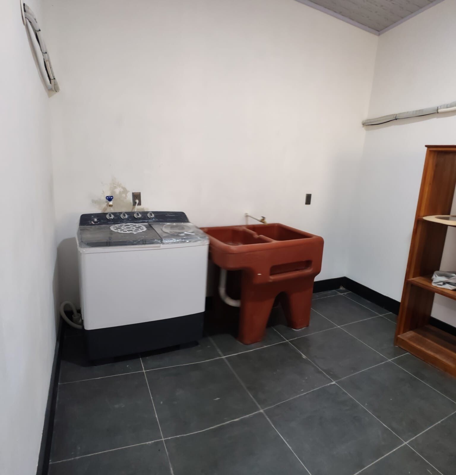 Ein kleines Zimmer mit Waschmaschine, einem roten Kunststoffhocker und einer Holzregaleinheit.