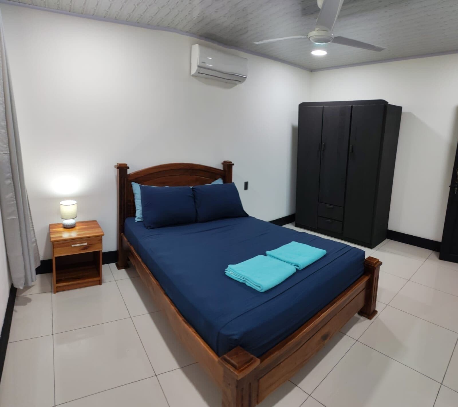 A neatly made bed with a wooden frame, a small bedside table with a lamp, a wardrobe, and an air conditioning unit on the wall.