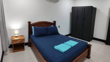 A neatly made bed with a wooden frame, a small bedside table with a lamp, a wardrobe, and an air conditioning unit on the wall.