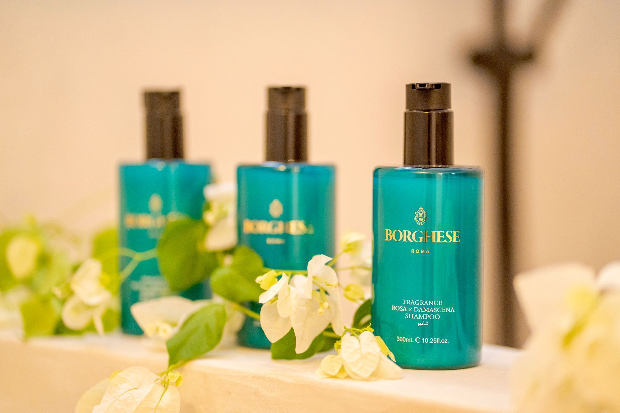 Three bottles of Borgheese fragrance shampoo on a shelf with white flowers.
