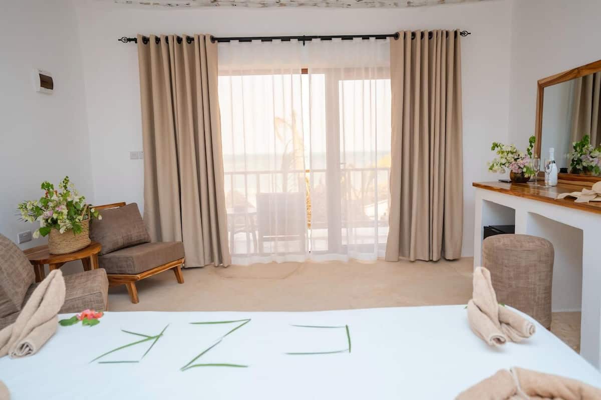 Une chambre d’hôtel comprenant un lit, deux fauteuils, une table d’appoint en bois avec un vase de fleurs, un bureau avec un miroir et un balcon avec des rideaux.