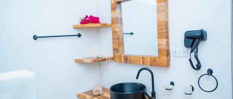 Une salle de bain avec un meuble-lavabo en bois, un lavabo noir, un miroir, une serviette, un sèche-cheveux et une petite étagère ornée de fleurs.