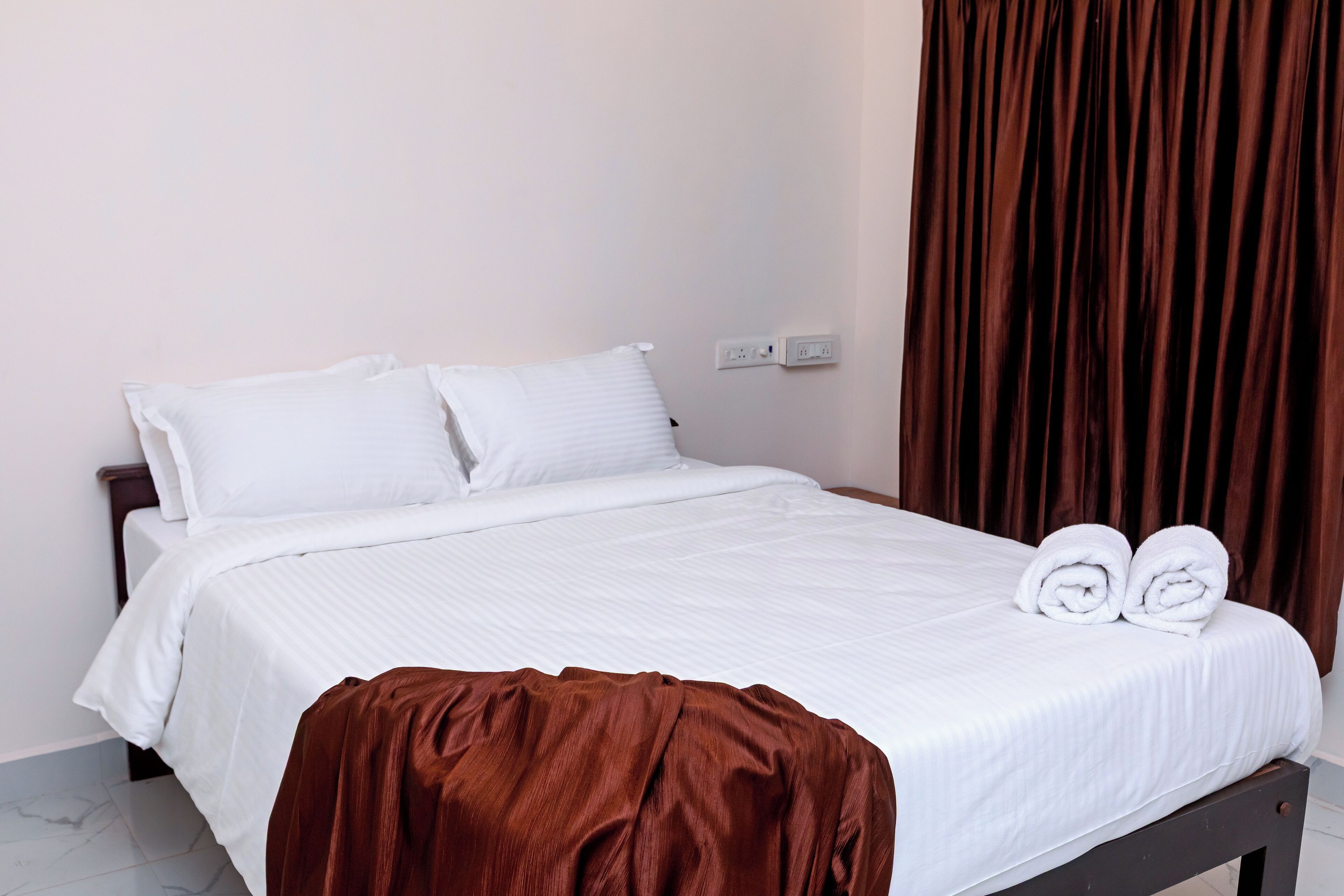 A neatly made bed with white linens and a brown throw blanket, a pair of rolled towels on the bed, and a dark wooden headboard.