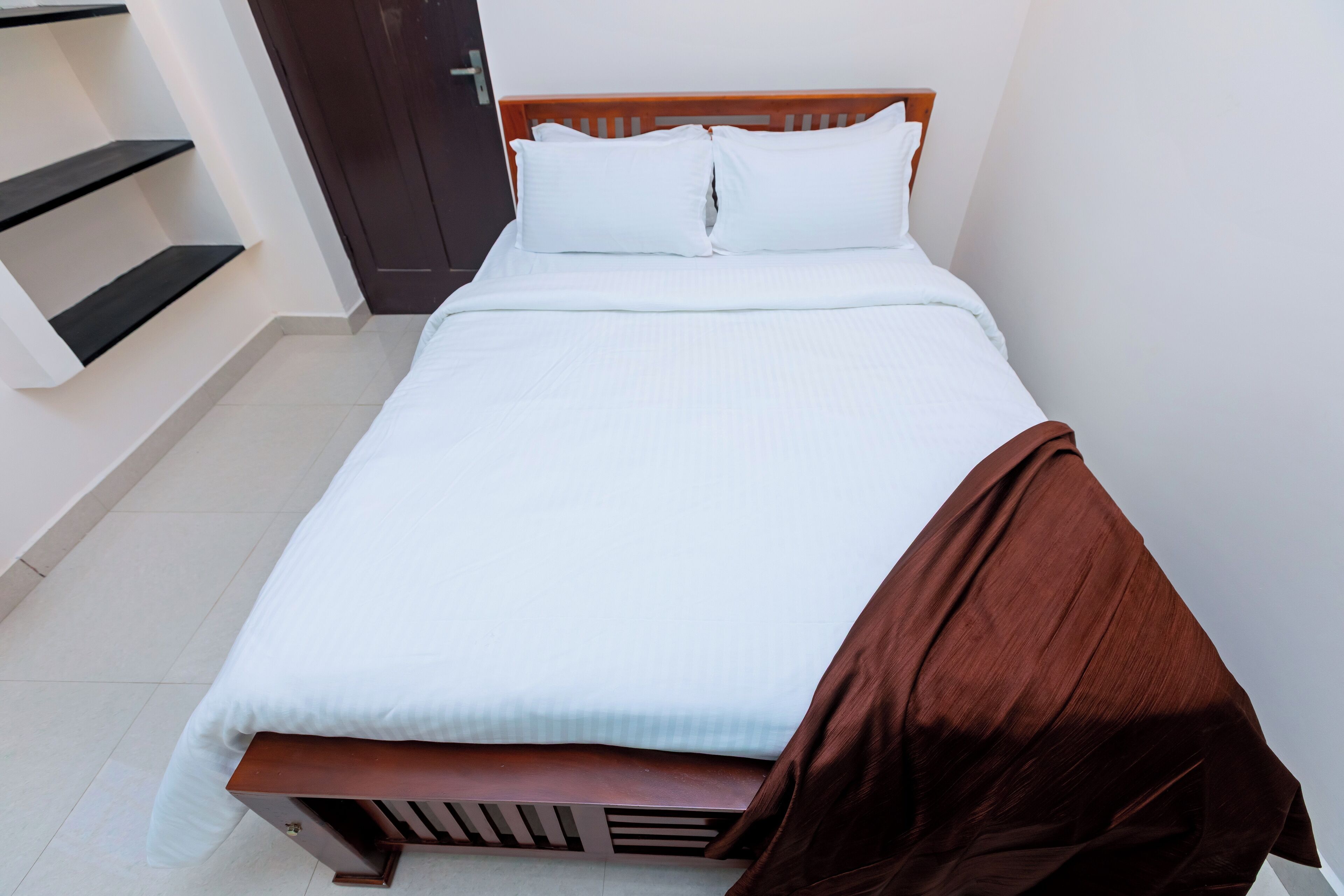 Una cama bien tendida con ropa blanca y una manta marrón, un cabecero de madera y una pared blanca con una puerta a la izquierda.
