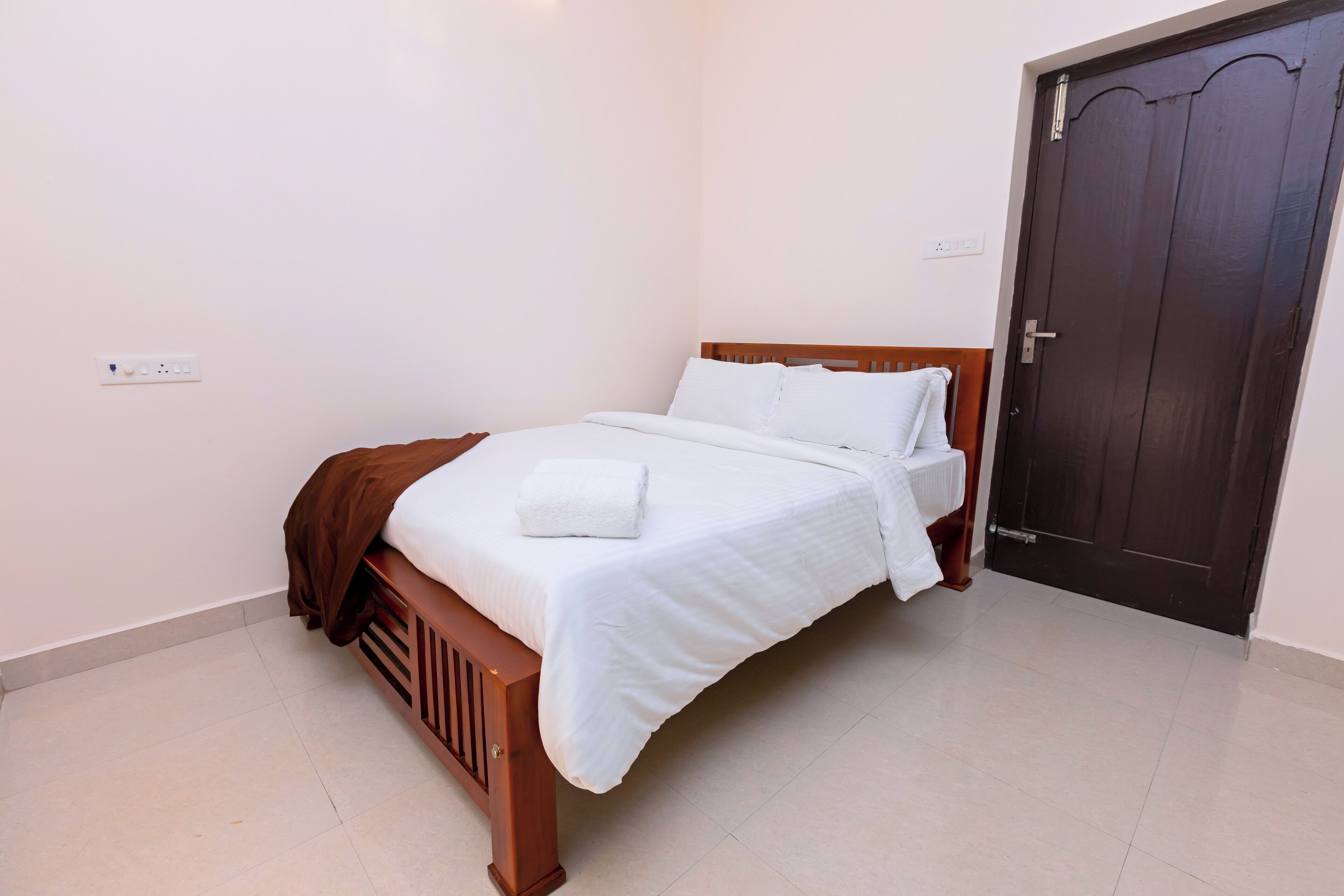 Una cama bien tendida con sábanas blancas y un cabecero marrón en una habitación con una puerta de madera oscura y paredes blancas.