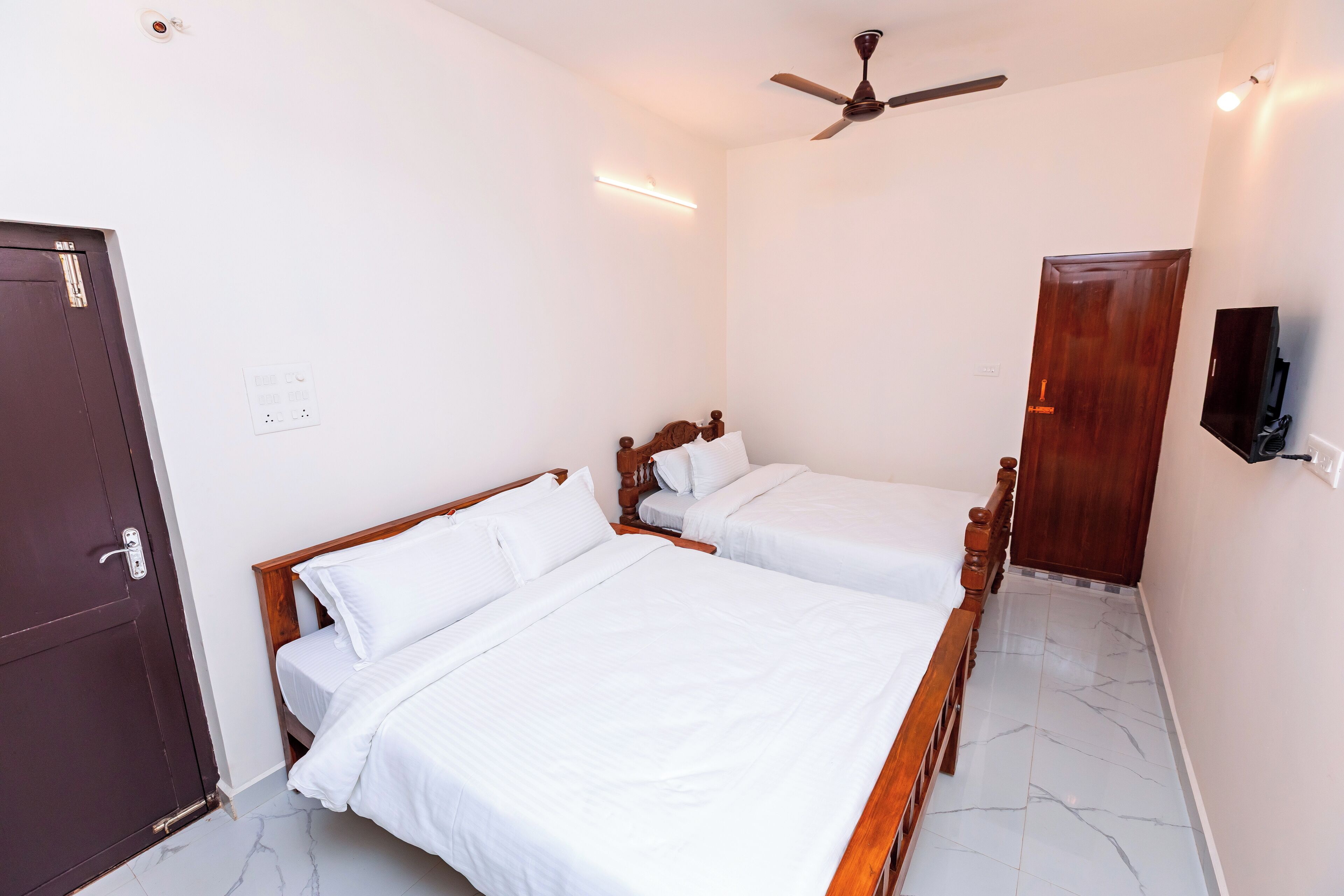 A hotel room with two single beds, a ceiling fan, a TV mounted on the wall, and a wooden door.