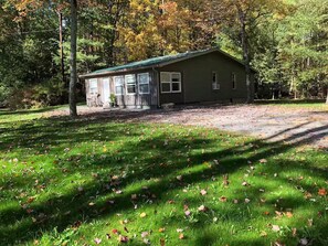 A small cabin with a green roof, surrounded by a grassy area with scattered leaves, and dense trees in the background.