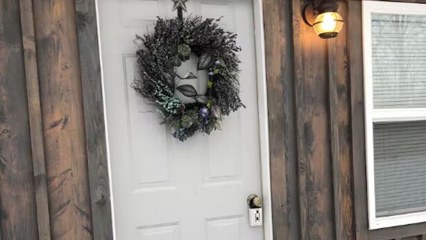 Una puerta blanca con un adorno festivo, un aplique de pared y una ventana con persianas.