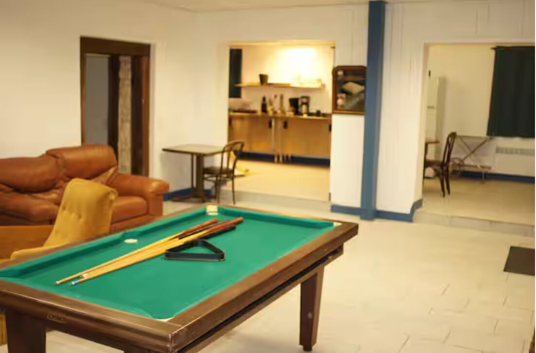 A pool table with pool cues, a brown leather sofa, and a small table with chairs in a room with white walls and a tiled floor.