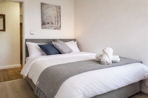 Een net opgemaakt bed met wit en grijs beddengoed, een grijze plaid en een opgerolde handdoek op het voeteneinde.